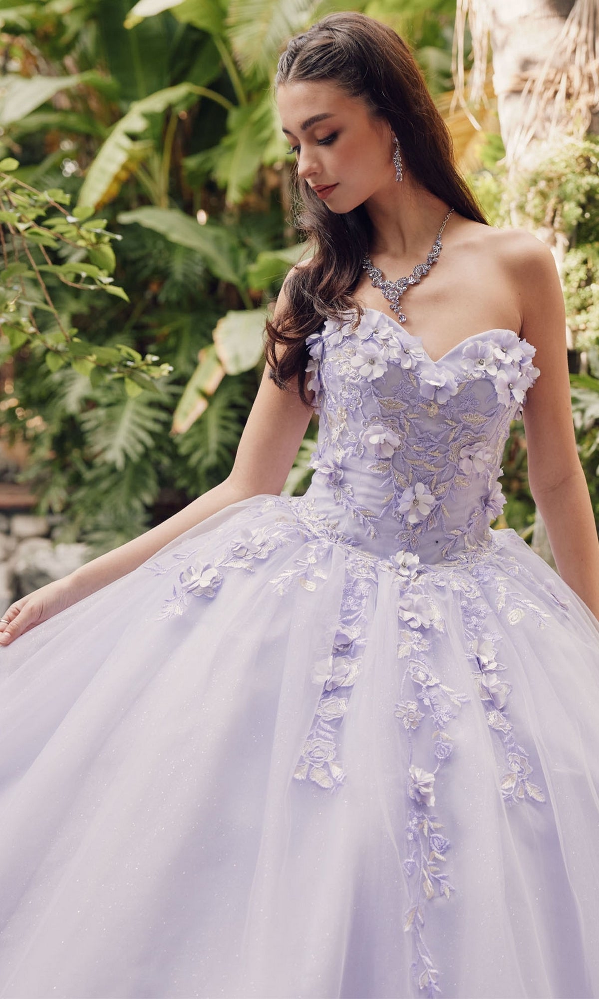 A woman in the Juliet 1435 Strapless Ball Gown Quinceañera Dress with floral appliqués stands outdoors holding her skirt, looking down, as lush green plants fill the background.